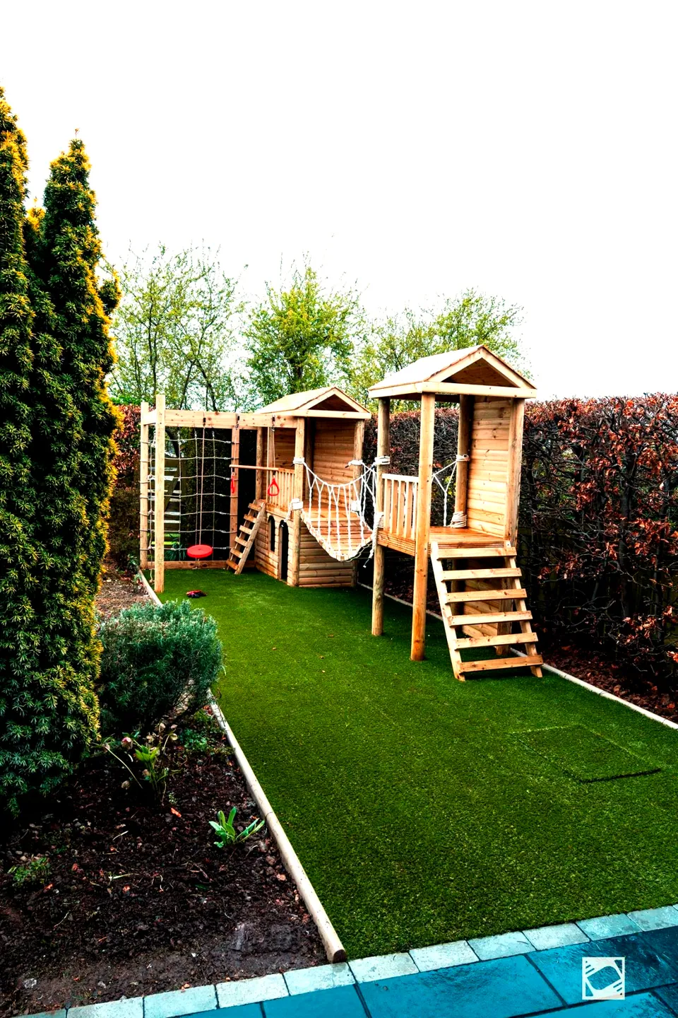 Timber playhouse with twin towers, a rope bridge and a climbing net on artificial grass
