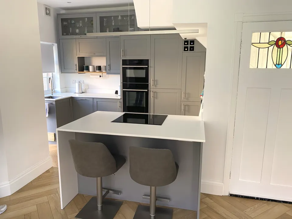 Grey shaker kitchen with a white breakfast island, induction hob and two bar stools on a herringbone floor