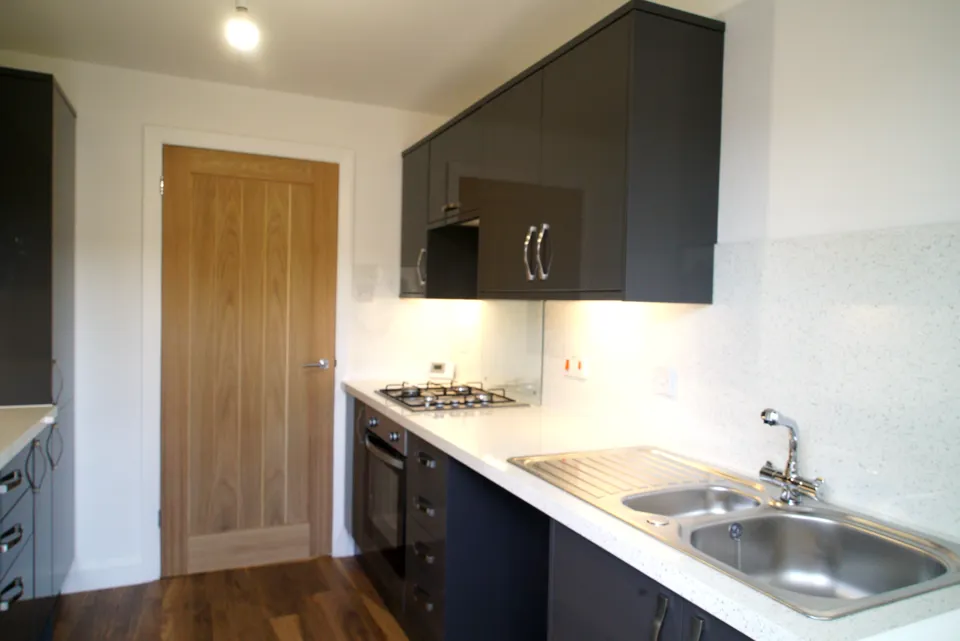 Black gloss galley kitchen with a white quartz worktop, stainless sink, gas hob and an oak door
