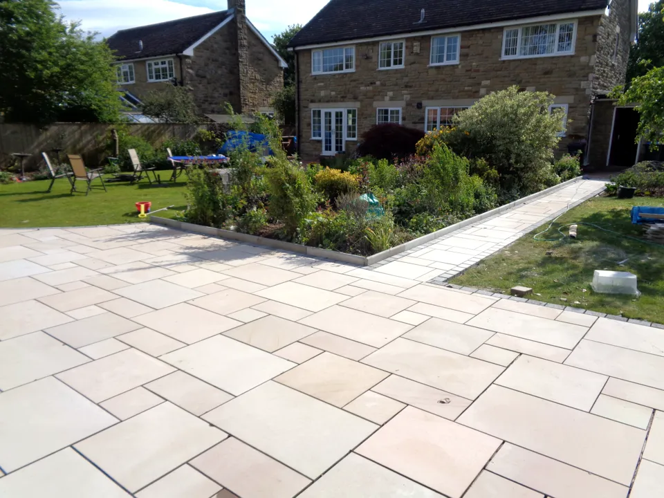 Finished sandstone patio laid in a random pattern, with planted borders and a lawn behind a stone house