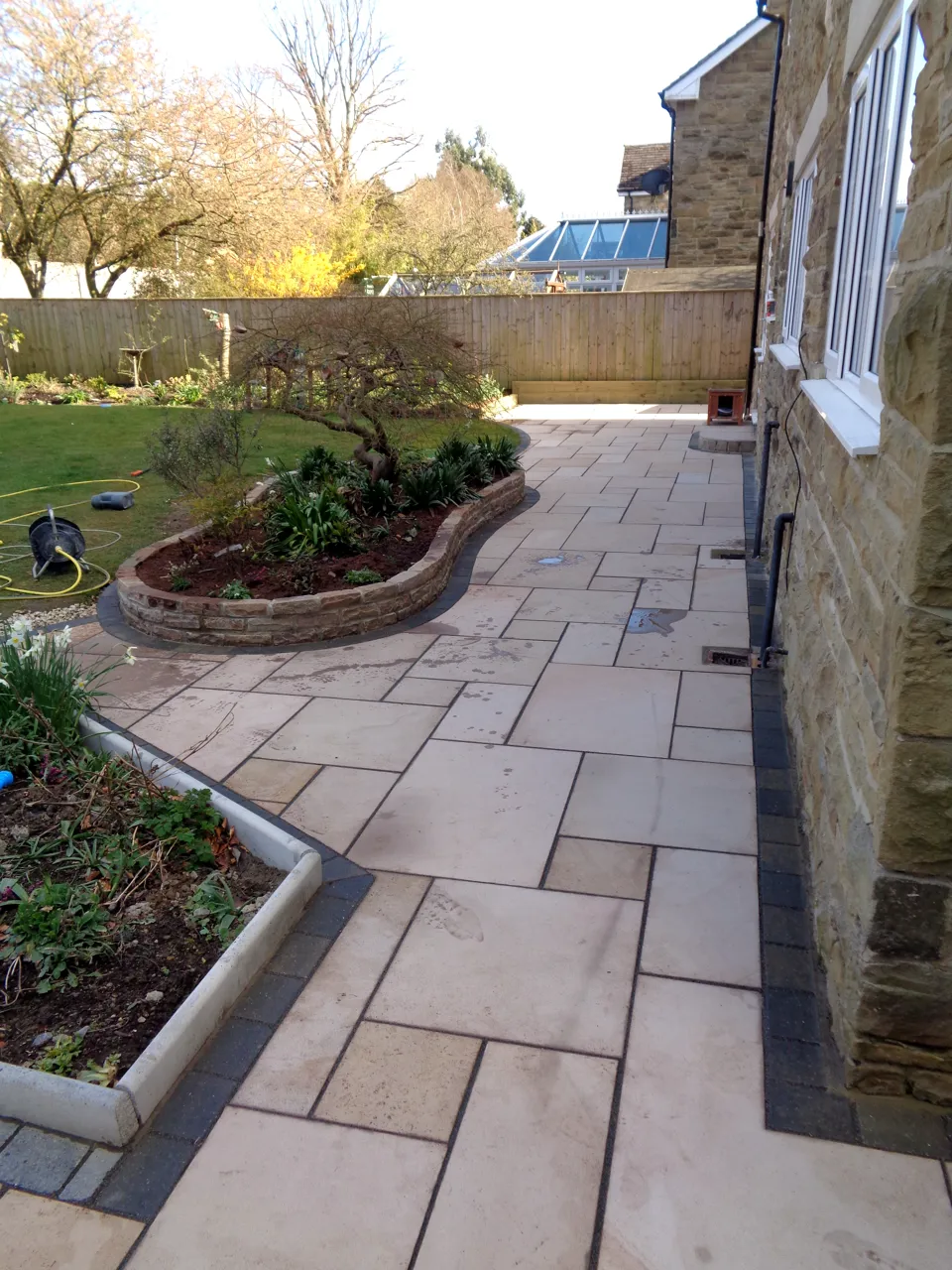 Sandstone patio curving past a raised bed along the side of a stone house