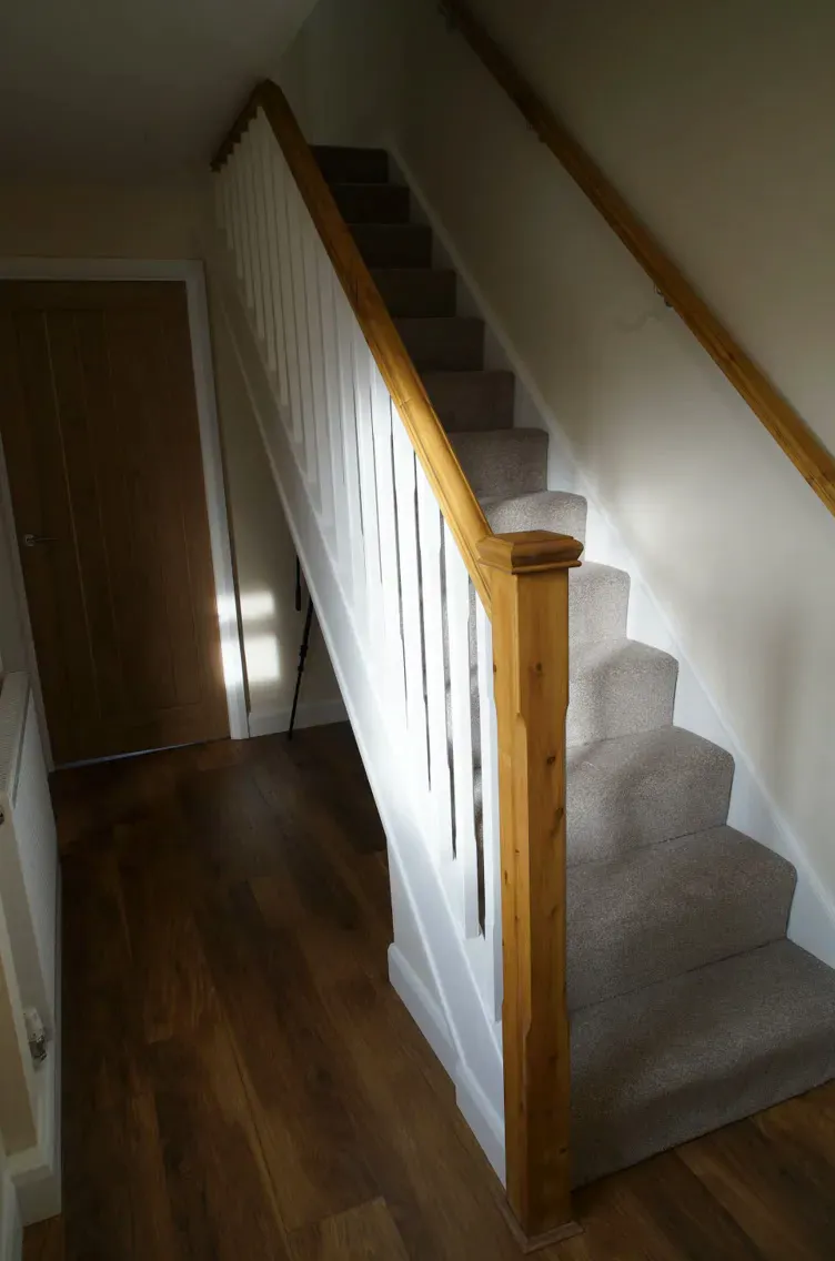 After: the same staircase with white spindles, a pine handrail and newel post, grey carpet and a wood-effect hall floor